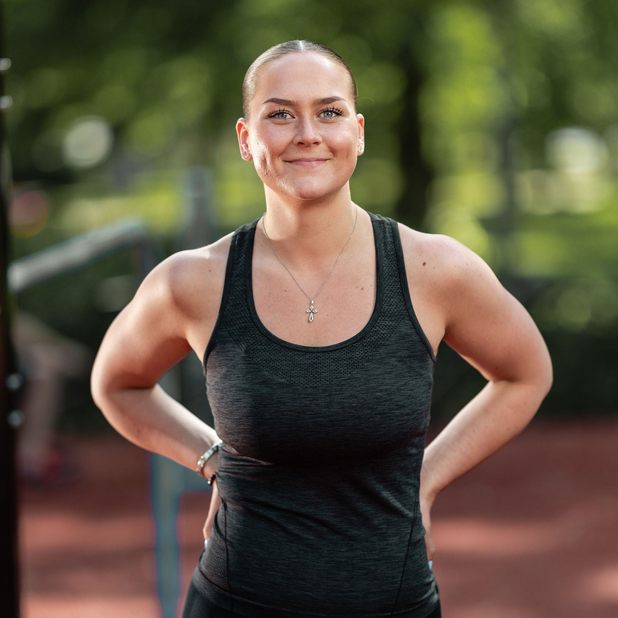 A woman standing outdoors on a park exercise court wearing a dark gray heathered athletic tank top and black leggings.